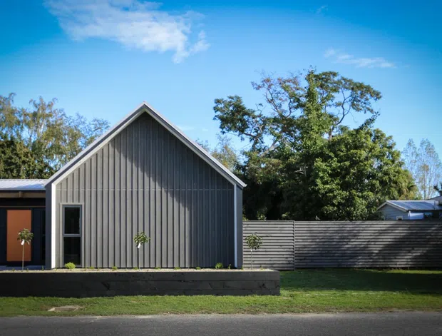 Street-facing view of a compact contemporary home with a prominent steep gabled volume clad in vertical grey timber boards, white edge trims, and a dark low horizontal wing. A small raised planter and lawn sit in front, with mature trees and neighbouring roofs behind.