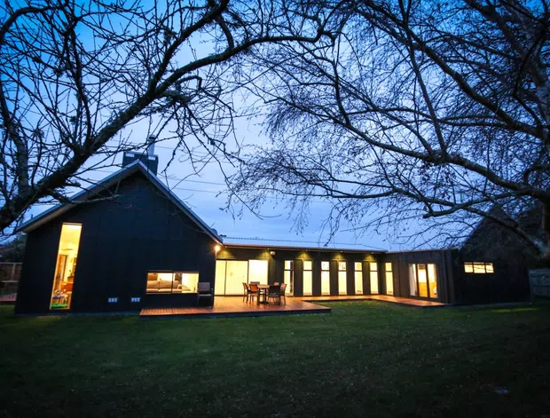 Exterior dusk view of a dark-clad, single-storey residential building arranged in an L-shaped form around a lawn. The home has a pitched gable-roofed wing connected to a long low-roofed wing with extensive warm-lit glazing and a timber deck or patio. Mature leafless trees frame the property.