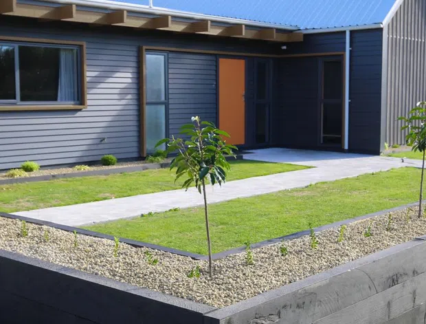 Single-storey contemporary residential house viewed from the front, with dark grey horizontal cladding, a steep gabled roof, timber-toned entry door, large windows, and a neat raised front garden bordered by a low concrete block retaining wall. A concrete path crosses the lawn to the recessed entrance.
