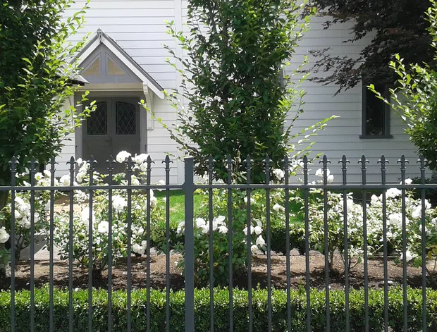 Front exterior view of a traditional white weatherboard house, partially screened by neatly clipped trees and a dark metal picket fence. The facade features a small gabled porch with decorative timber detailing, narrow windows, and a landscaped garden with white flowering shrubs.