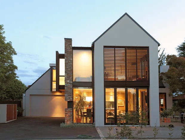 Front exterior view of a contemporary multi-level residence with a prominent steep gabled facade, smooth grey rendered walls, extensive glazing, warm timber horizontal sunshades, and a tall stone-clad vertical element. The house is set among mature landscaping and includes an attached garage.