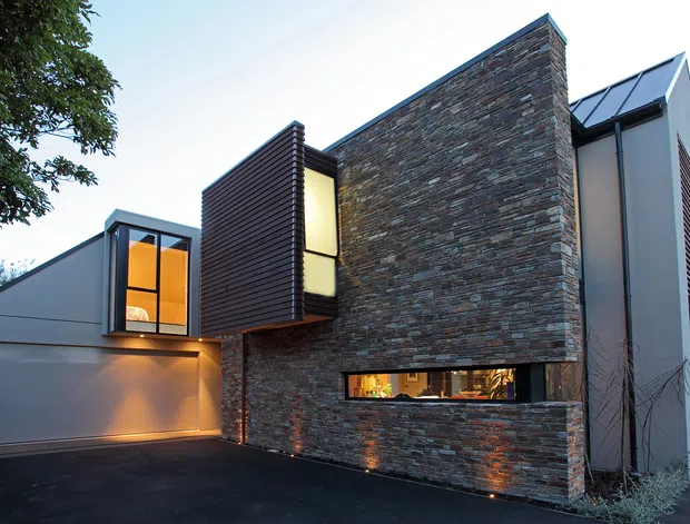 Contemporary multi-level house exterior at dusk, featuring a dramatic angular form, dark horizontal cladding, extensive stacked-stone wall cladding, narrow illuminated windows, a large garage door, and a steep metal roof element. The home is viewed from the driveway with subtle architectural lighting highlighting the facade.
