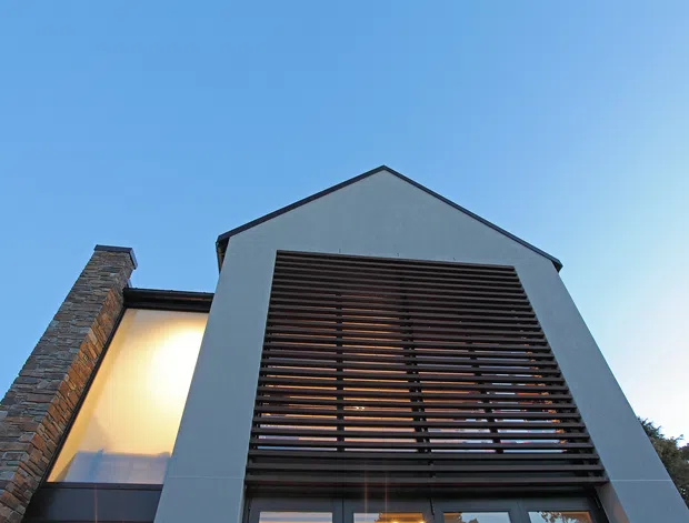Low-angle exterior view of a contemporary multi-storey building facade beneath a clear blue sky. The central gabled volume has a smooth grey rendered finish and a large dark horizontal louvred screen over glazing, while the adjoining section features stacked stone cladding and illuminated glazing.