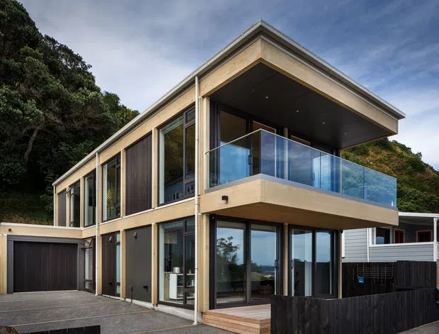 Exterior view of a contemporary two-storey residence on a sloping, densely vegetated site. The home has a strong projecting upper-floor balcony with clear glass balustrading, extensive floor-to-ceiling glazing, pale timber or timber-look cladding, dark vertical wall panels, a broad flat or very low-pitched roof, and an integrated single-car garage. A neighbouring house is visible to the right.