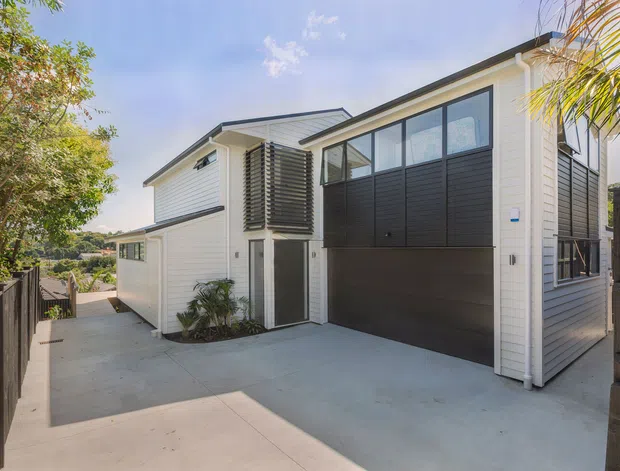 Exterior view of a contemporary multi-level home with white horizontal cladding, dark contrasting garage door and upper wall panels, large black-framed windows, and a broad concrete driveway or forecourt. Timber fencing and planted areas frame the property.