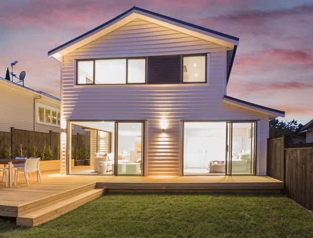 Twilight exterior view of a compact two-storey suburban home with a prominent gabled roof, horizontal weatherboard cladding, extensive glazing, and a timber deck with outdoor dining. Warm interior lighting is visible through the ground-floor windows and doors, with neighbouring houses and fencing surrounding the property.