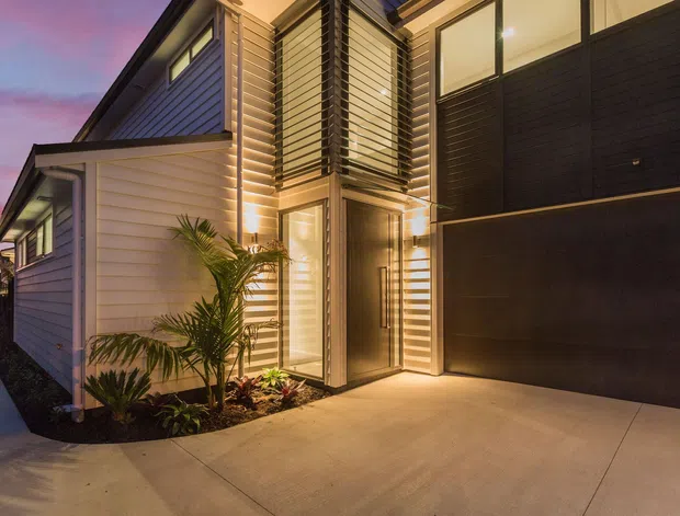 Contemporary multi-level house exterior at dusk, featuring light horizontal weatherboard cladding, dark vertical cladding panels, a tall glazed entry, horizontal sun-shading screens, warm exterior lighting, and a broad concrete driveway. Tropical planting frames the entry.