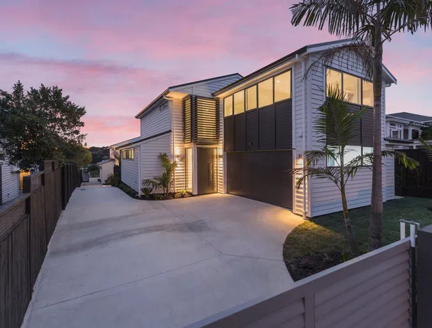 Contemporary two-storey residential home viewed from the driveway at dusk. The facade combines white horizontal cladding, dark charcoal panels, warm exterior lighting, large upper-level glazing, and a broad concrete driveway. Palm trees and neighboring homes frame the suburban setting.