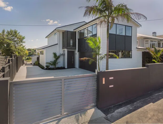A contemporary multi-level residential home viewed from the street, featuring white horizontal cladding, dark charcoal wall panels, large black-framed windows, and a low dark boundary wall with a matching sliding garage gate. Palm trees and a narrow paved entry path frame the modern facade.