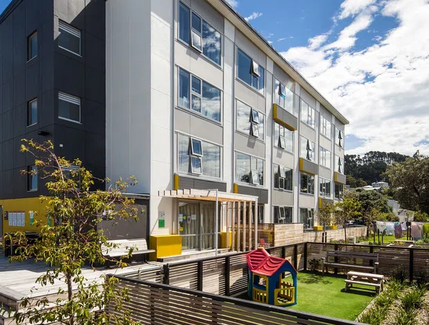 Exterior view of a contemporary multi-storey apartment or residential building, with a tall white and dark grey panelled facade, rows of windows, projecting balconies, and a covered entrance. The landscaped foreground includes timber privacy fencing, planted garden beds, a communal deck, artificial turf, and children's play equipment. Bright sunny conditions and a blue sky are visible.