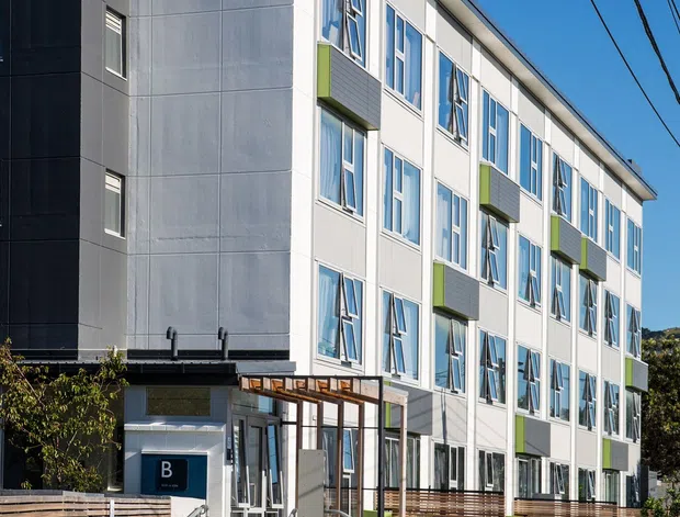 Exterior view of a contemporary multi-storey building with a long rectilinear form, pale grey and white facade panels, extensive vertical windows, and projecting lime-green balcony or privacy-panel accents. The entrance has a covered timber pergola, horizontal timber fencing, accessible ramping, landscaped planting, and a visible blue 'B' sign. Overhead utility wires cross the clear blue sky.