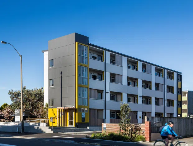 Street-level exterior photograph of a contemporary multi-storey apartment building. The building has a light grey rendered or panelled facade, strong vertical yellow accents around a glazed stair or lobby volume, balconies across multiple levels, dark window and railing elements, and a recessed ground-level entrance. A street, low boundary wall, landscaping, and a cyclist are visible in the foreground.