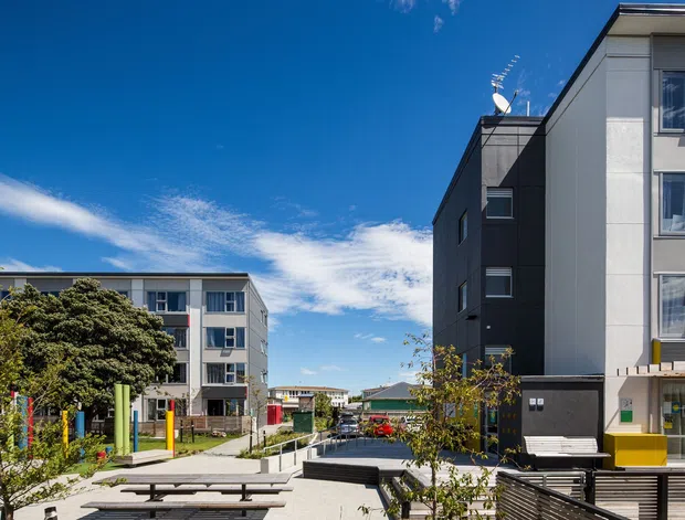 Exterior view of a contemporary multi-storey residential apartment development. The image shows several compact apartment buildings arranged around a landscaped pedestrian courtyard, with rendered white and dark grey facades, flat roofs, regular windows, paved walkways, timber-look seating or screening, and a bright blue sky.