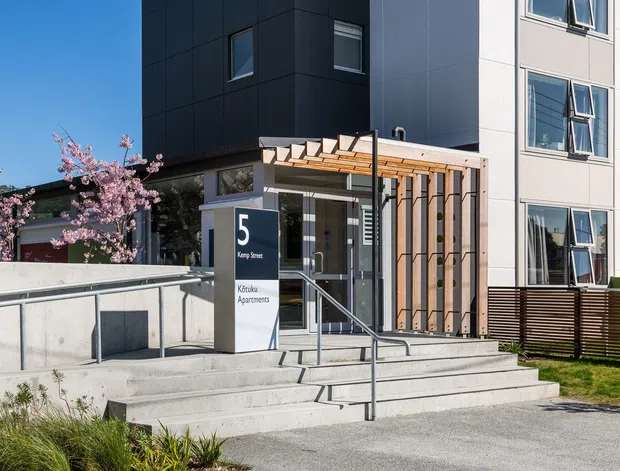 Exterior view of a contemporary multi-storey apartment building entrance. The facade combines dark metal cladding with light rendered or panelled walls, large rectangular windows, concrete stairs and accessible ramping, and a timber pergola-style canopy over the entry. A sign identifies the building as 5 Kemp Street and Kaitoke Apartments. Landscaping and a flowering tree frame the entrance.