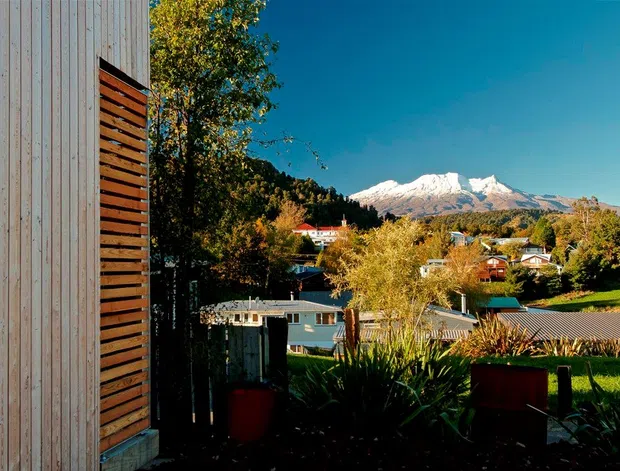 Exterior architectural view of a contemporary building with vertically clad light timber walls and a dark horizontal timber privacy screen, overlooking a suburban or semi-rural landscape with a prominent snow-capped mountain backdrop. The image is brightly lit with strong natural contrast and autumnal vegetation.