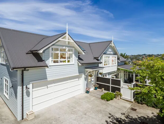 Exterior view of a multi-level suburban residence with pale blue-grey weatherboard cladding, steep dark metal gabled roofs, decorative gable detailing, dormer-style projecting windows, white trim, and an integrated double garage. A paved driveway, entry gate, landscaping, and neighbouring hillside homes are visible.