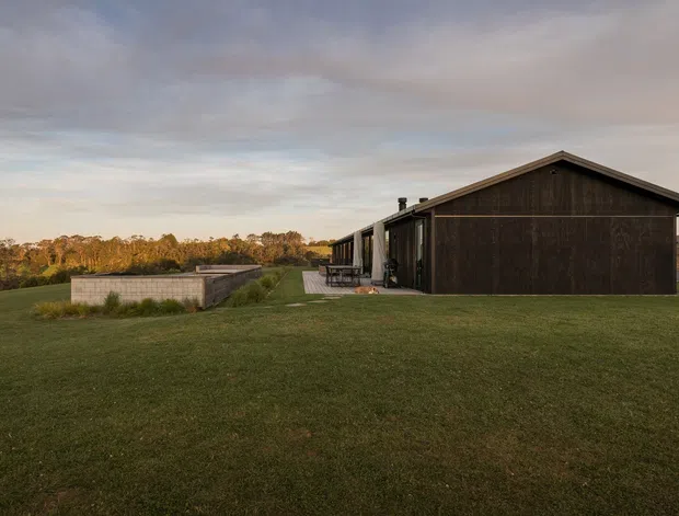 Single-storey dark-clad rural house set within a broad mown lawn. The simple gabled form has dark timber or wood-look wall cladding, a projecting roof edge, and a recessed glazed side elevation with a small deck or entry platform. Low masonry landscape walls and open rural surroundings are visible, with warm late-day light.