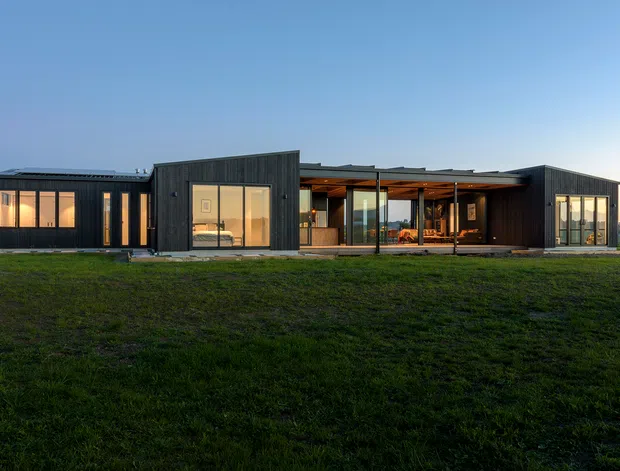 A single-storey contemporary residence viewed from across a broad lawn at dusk. The long, low building has several dark vertical-clad volumes connected by extensive floor-to-ceiling glazing and a recessed, covered central outdoor living area. Warm interior lighting contrasts with the dark facade, while the flat or very low-pitched roofline reinforces the horizontal modern form.