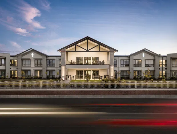 Symmetrical multi-storey institutional or commercial building photographed from across a roadway at dusk. The light-coloured facade features repeated gabled roof sections, dark-framed windows, illuminated interiors, landscaped frontage, and a prominent central entrance pavilion with a balcony, exposed triangular roof framing, and large supporting columns. Long-exposure vehicle light trails run across the foreground.