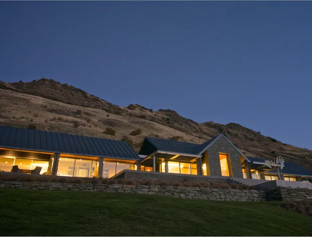 Exterior dusk view of a low-slung contemporary residential building set against a dry, rocky hillside. The home features multiple pitched roof volumes with dark metal roofing, extensive warm-lit glazing, stone-clad walls and retaining walls, and a gently landscaped foreground.