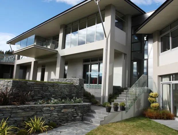 Exterior view of a contemporary multi-level residence with broad low-pitched roof planes, white rendered walls, extensive floor-to-ceiling glazing, projecting balconies, and a tall glazed entry stairwell. Layered dark stone retaining walls and slate-like paving form terraced landscaping in the foreground.