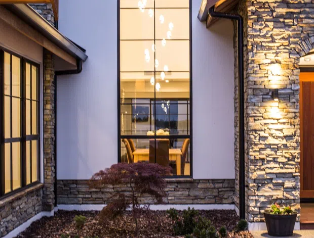 Exterior view of a contemporary residential entry courtyard at dusk, framed by wings with dark roof edges, gutters, stacked natural-stone veneer walls, and a very tall arched window with dark mullions. Warm interior lighting and a visible dining area show through the glazing, while low landscaping and a concrete path lead toward a timber entry door.