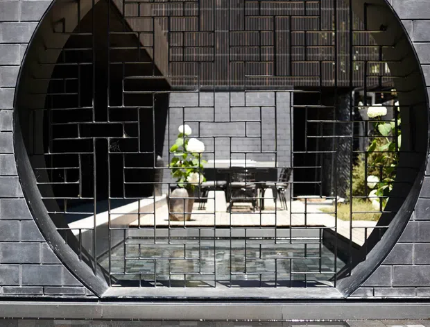 A striking contemporary architectural facade clad in dark grey brick, featuring a large circular opening with a black geometric metal grille and reflective glazing. Through the opening, a bright courtyard or interior space with plants, seating and shelving is visible. A shallow reflective pool with dark stone paving occupies the foreground, creating a dramatic layered composition.