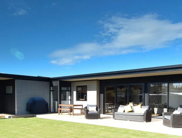 Single-storey contemporary residence viewed from the lawn, with a long low-profile form and shallow roofline. Dark exterior cladding and extensive floor-to-ceiling glazing frame a sheltered outdoor entertaining area furnished with lounge seating and a dining table. The building opens onto a broad lawn under a clear blue sky.