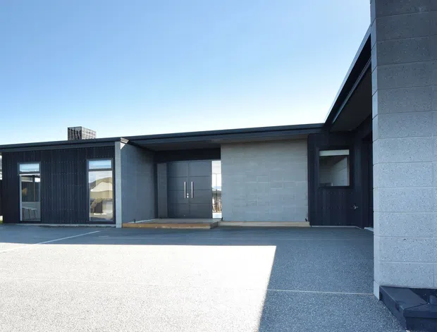 Contemporary single-storey building exterior viewed from a broad paved forecourt. The facade combines dark vertical cladding, pale concrete masonry walls, large glazed openings, a recessed entry, and a low-profile roofline. A taller pale masonry volume projects at the right, with minimal planting and strong sunlight creating crisp shadows.