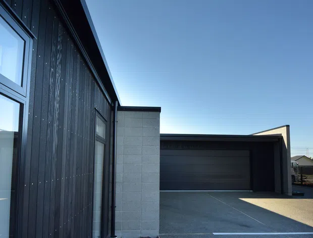 Exterior view of a contemporary dark-clad building with large glazed openings and a prominent attached double garage. The facade combines vertical dark cladding, pale masonry/concrete blockwork, broad garage doors, and a paved driveway under a clear sky.