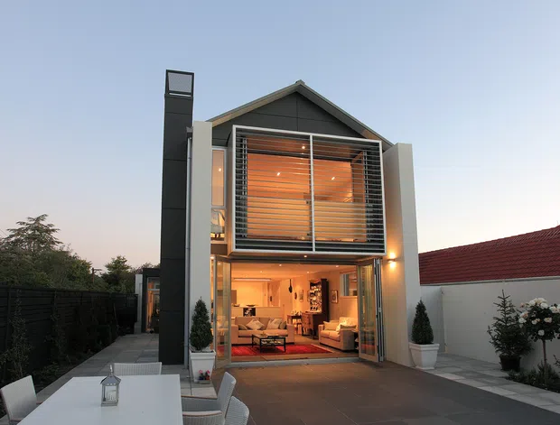 A contemporary multi-level house viewed from the front at dusk, featuring a steep gabled facade, extensive floor-to-ceiling glazing, a tall dark vertical chimney or wall element, and a warm timber-lined upper-level interior visible behind horizontal screening. The ground floor opens toward a paved courtyard or driveway and reveals a warmly lit living area. A neighbouring red-tiled roof and landscaped planting are visible at the sides.