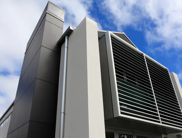 Low-angle exterior view of a contemporary multi-storey building with sharp geometric forms, dark panel cladding, pale rendered or panelled vertical elements, and a prominent projecting facade screened with horizontal metal louvres. A partly cloudy blue sky is visible behind the structure.