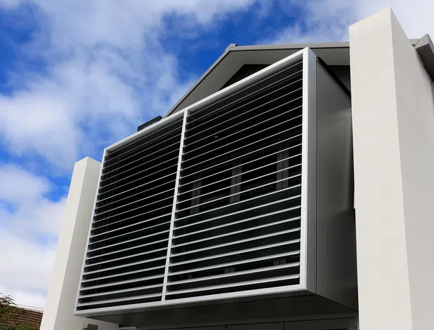 Low-angle exterior view of a contemporary multi-level building facade featuring a large projecting louvred screen, white rendered wall planes, dark glazing, and a flat or shallow-pitched roof form beneath a partly cloudy sky.