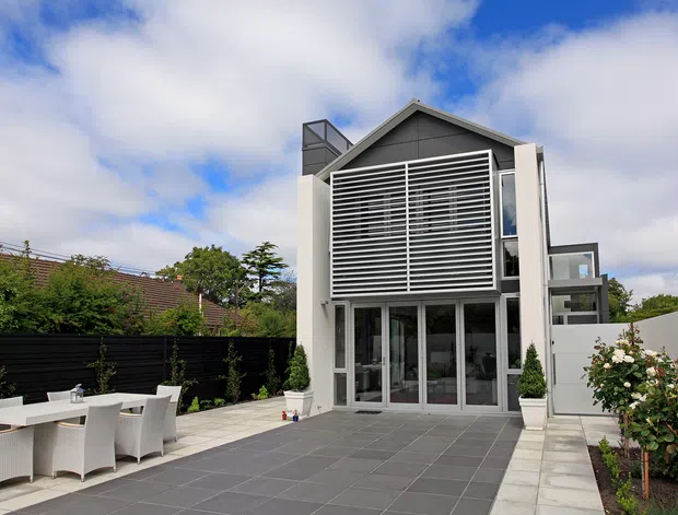 Contemporary multi-level residential exterior viewed from a paved courtyard or terrace. The house has a steep gabled roof, dark grey upper cladding, white rendered framing, extensive glazing, and a prominent horizontal louvred screen across the upper facade. Outdoor dining furniture, planter boxes, fencing, and landscaped borders are visible in the foreground.