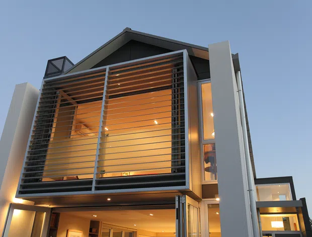 Upward-looking exterior view of a contemporary multi-level residence with a pronounced gabled roof, large glazed openings, and a projecting upper-level balcony or sunroom screened by horizontal timber louvres. White vertical facade elements, dark framing, warm interior lighting, and recessed exterior lights create a modern architectural presentation at dusk.