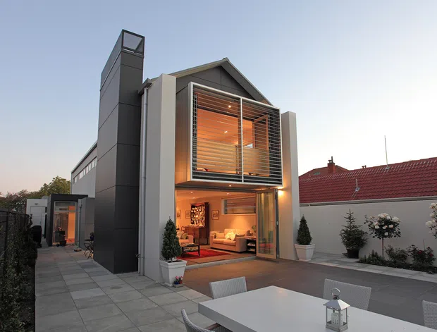 Contemporary multi-level residential extension viewed from a paved courtyard, with a steep gabled roof, dark vertical chimney-like structure, large glazed openings, and an upper-level timber louvred screen. Warm interior lighting reveals a living area through the glass, while a white outdoor dining table and chairs occupy the foreground. The property is enclosed by rendered walls and landscaped with potted and planted shrubs.