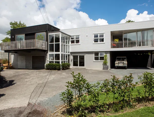 Exterior view of a contemporary multi-storey residential house with a broad horizontal composition. The home combines light grey horizontal cladding, dark upper-level cladding, extensive glazing, projecting balconies with timber balustrades, a glazed stairwell or entry volume, and an open garage beneath the right side. A concrete driveway and landscaped planting occupy the foreground.