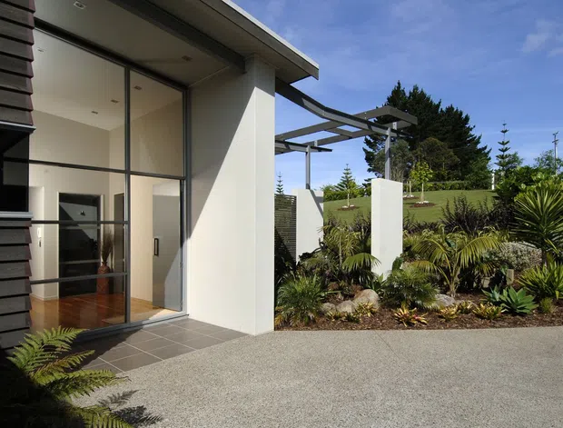 Modern residential exterior viewed from a landscaped entry or driveway, featuring dark horizontal cladding, white rendered walls, expansive floor-to-ceiling glazing, a projecting flat roof, and a lightweight pergola structure. Tropical and subtropical planting surrounds the paved approach.