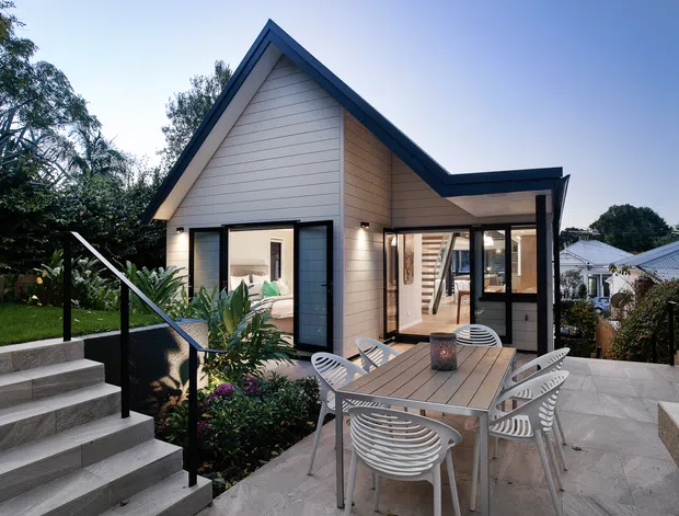 Exterior view of a compact contemporary gable-roof home or extension, with pale horizontal weatherboard cladding, dark trim, extensive glazing, and a paved outdoor dining terrace. A stairway with black metal handrails leads up through landscaped garden beds to the terrace. The image is taken from outside and the building facade and terrace dominate the frame; a softly furnished interior is visible through the windows.