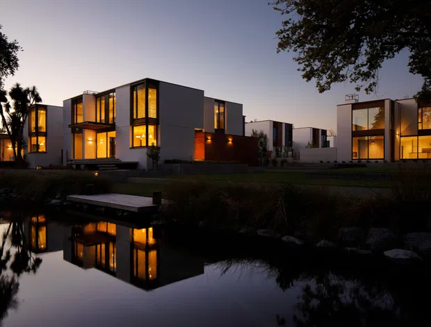 A dusk exterior view of a modern multi-building residential development beside a reflective pond or landscaped waterway. The buildings feature crisp cubic forms, flat roofs, dark and light facade planes, extensive floor-to-ceiling glazing, warm interior lighting, and timber-toned feature panels. A small jetty extends over the water in the foreground, with mature trees framing the scene.