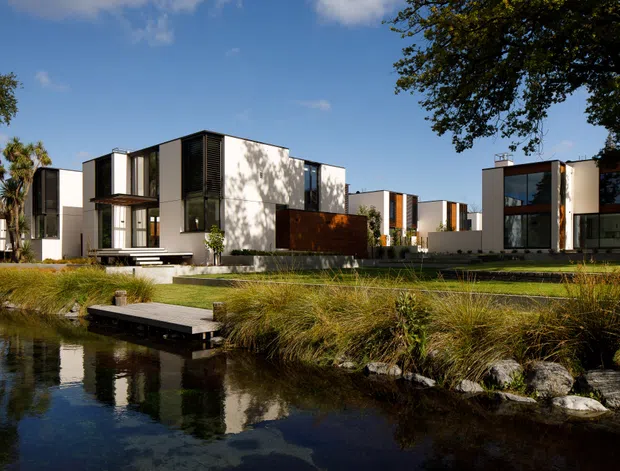 A contemporary multi-building residential development viewed across a landscaped pond. The buildings feature crisp white cubic volumes, dark vertical louvres or cladding, extensive glazing, warm timber accents, and flat roofs. Native-style grasses, rock edging, and a small timber jetty frame the foreground, creating a landscaped waterside setting.
