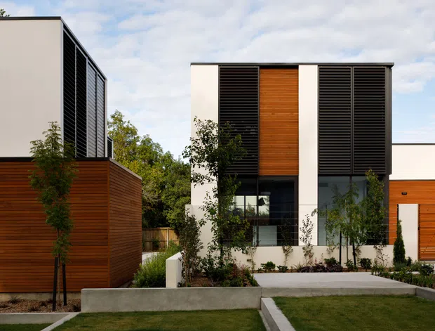 Contemporary multi-level residential facade with crisp white rendered walls, extensive dark horizontal louvres, and warm timber-clad sections. The composition includes landscaped front gardens, concrete retaining or garden edges, and flat roof forms under a partly cloudy sky.