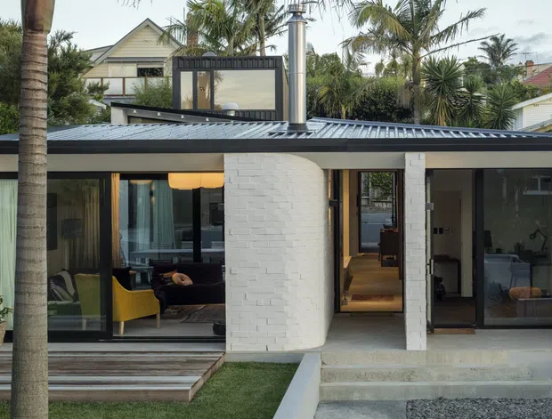 Exterior view of a low, contemporary single-storey residence with a flat roof, dark metal roof and window framing, white painted brick walls, extensive glazing, and a rooftop terrace or upper-level pavilion. Palm trees and lush landscaping surround the home, with interior living spaces visible through the windows.
