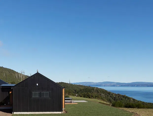 Exterior view of a contemporary pair of gabled buildings on a sloping rural/coastal site. The foreground structure has warm timber cladding and a large garage door, while the darker adjacent building has vertical dark cladding and a simple pitched roof. A broad body of water and wooded headland are visible beyond.