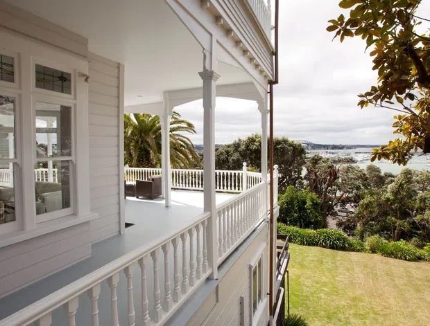 Exterior view of a light-painted weatherboard house with ornate white trim, decorative sash-style windows, and a covered two-level veranda with turned balustrades and columns. The property overlooks a landscaped garden and a visible coastal bay or waterfront beyond.