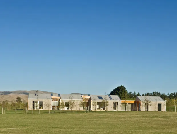 A low, elongated rural property viewed from across a broad lawn, composed of several connected gabled volumes with pale masonry or rendered walls, dark pitched roofs, extensive glazing, and warm timber accents. Fenced paddocks, trees, and dry rolling hills establish a rural setting under a clear blue sky.