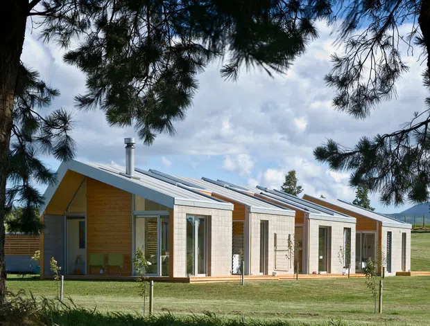 Exterior view of a low-profile contemporary residential building composed of several connected gabled volumes. The facade combines warm timber cladding with pale masonry or rendered panels, extensive glazing, and repeated sloping roof planes with skylight-like raised sections. The building sits in a landscaped grassy setting, framed by mature trees, with distant open countryside visible beyond.