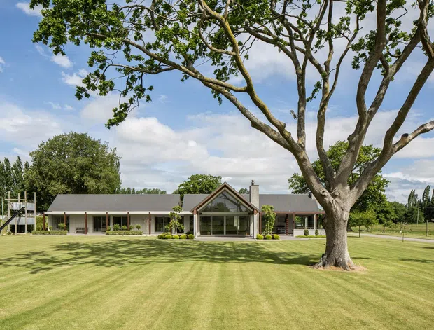 A single-storey residential property viewed from a broad, manicured lawn. The house has a long low-profile form with dark grey pitched roofing, light exterior walls, extensive glazing, and a prominent glazed gabled entrance or central pavilion. Mature trees frame the foreground, with open landscaped grounds surrounding the building.