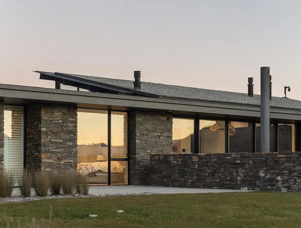 Exterior view of a low, single-storey contemporary house at dusk. The building has extensive floor-to-ceiling glazing, dark stone masonry walls and a low-pitched roof with multiple chimney or flue elements. A projecting covered terrace or courtyard wall runs along the facade, with landscaped lawn and grasses in the foreground.