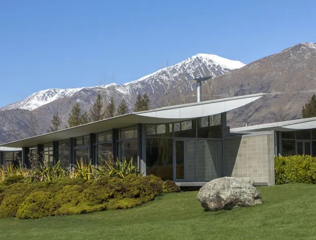 Single-storey modernist house set in a landscaped garden, with long horizontal wings, broad low-pitched projecting roof planes, extensive dark-framed glazing, and pale masonry walls. Snow-capped rugged mountains form the visible backdrop, while shrubs, lawn, and a sculptural boulder surround the building.